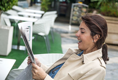 Smiling woman reading newspaper while sitting at table