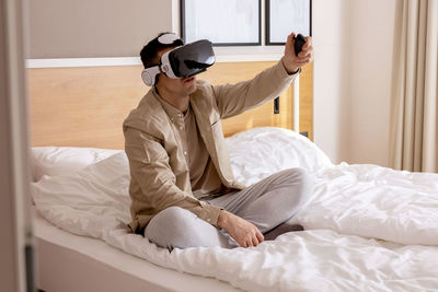 Young caucasian man sitting on bed at home with vr headset and playing interactive video game