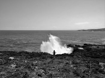 Rear view of woman looking at sea while standing on rocky beach against sky