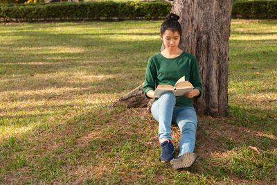 Full length of young woman reading book while leaning on tree at park