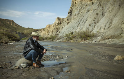 Adult man in cowboy hat sitting on rock along river flows in desert. almeria, spain