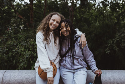 Portrait of smiling female friends with arm around sitting on retaining wall