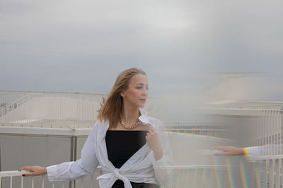 Portrait of beautiful young woman standing on railing against sky