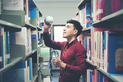 Young man holding globe amidst bookshelves in library