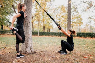 Happy friends exercising with straps while standing by tree