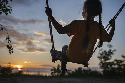 Rear view of girl sitting on swing during sunset