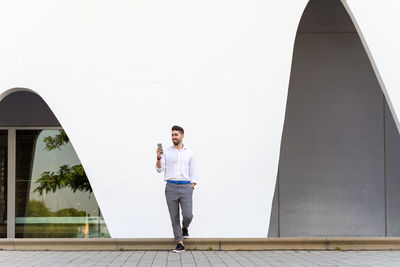 Young bearded man leaning against a white wall using the mobile phone