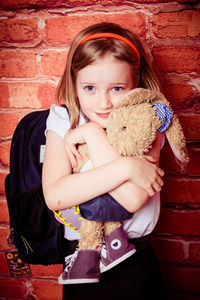 Portrait of schoolgirl embracing stuffed toy against brick wall