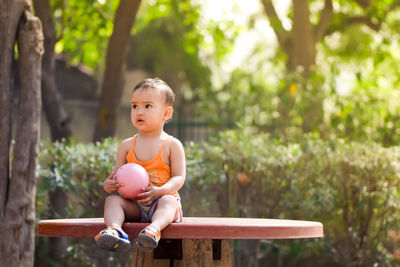 Full length of girl with ball looking away while sitting on bench at public park