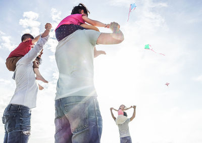 Low angle view of fathers carrying their children on shoulders while standing against sky during sunny day