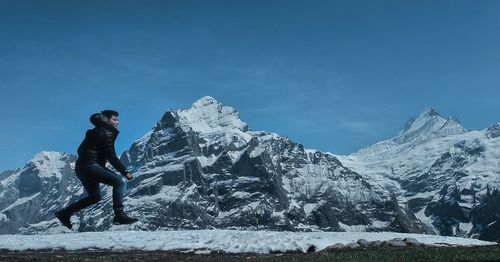 Side view of young man jumping on snow covered field against blue sky