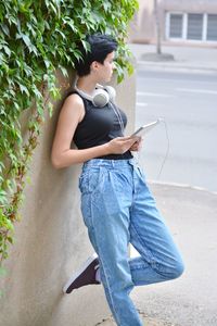 Thoughtful young woman holding digital tablet looking away while standing against wall