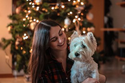 Smiling woman with dog against christmas tree at home