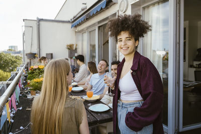 Portrait of smiling young woman with hand in pocket holding wineglass while celebrating dinner party with friends in bal