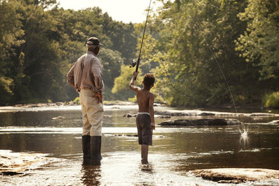 Rear view of boy fishing by grandfather standing by lake