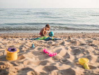 Side view of little girl sitting on haunches and playing with sand on sandy shore near waving sea