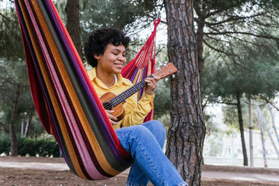 Smiling young ethnic female in bright shirt lounging in park hammock and playing ukulele