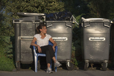 Young woman sitting on chair by wheeled garbage cans