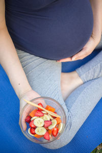 High angle view of pregnant woman holding bowl with fruits while sitting on exercise mat