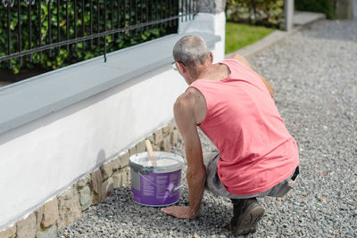 A middle-aged man paints a fence with white paint with a brush, repairs the damaged surface, photo