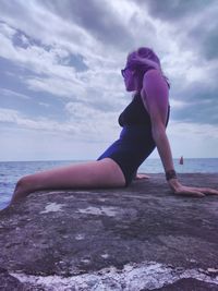 Side view of mid adult woman in swimwear relaxing at beach against cloudy sky