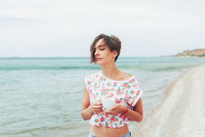 Woman holding coffee cup while standing against sea