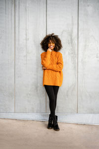 Full length of smiling ethnic woman with curly afro hair wearing vivid orange sweater looking at camera against gray wall