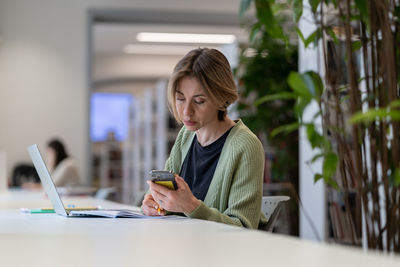 Focused female university teacher using laptop while sitting in quiet cozy library