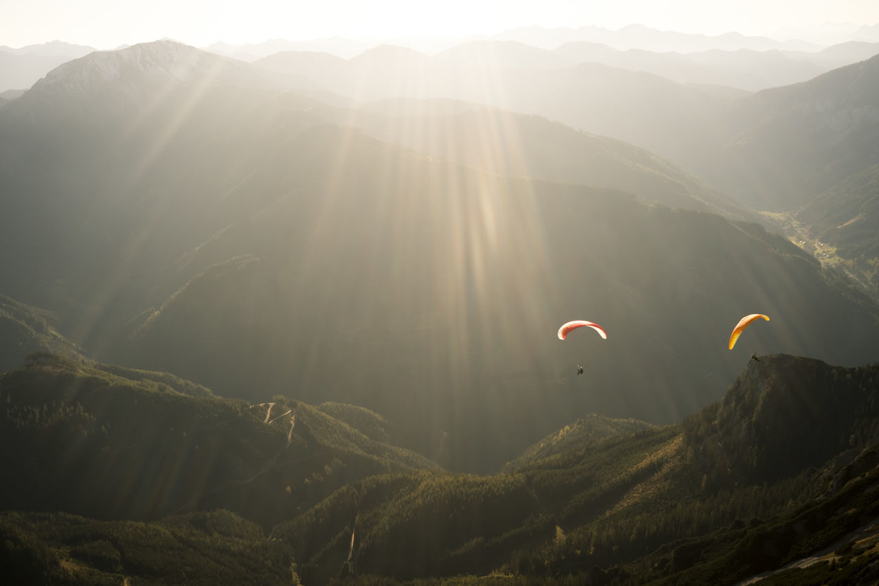 mountain, adventure, beauty in nature, sunlight, mountain range, parachute, sky, scenics - nature, sunbeam, paragliding, extreme sports, nature, tranquility, lens flare, tranquil scene, leisure activity, sun, environment, day, sport, freedom, outdoors, bright