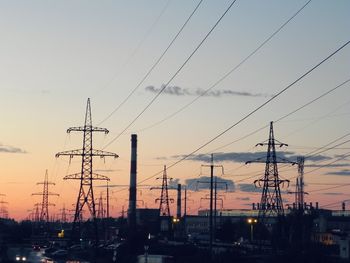 Electricity pylons amidst cityscape against sky during sunset