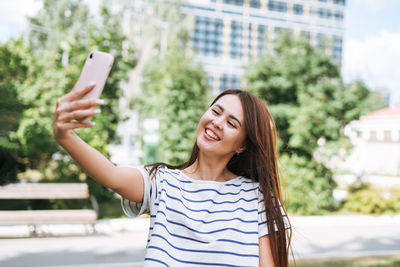 Portrait of young woman student with long hair taking selfie with mobile phone in city park