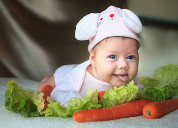 Portrait of cute baby girl wearing bunny hat while lying with carrots and lettuce on bed