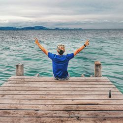 Rear view of man gesturing peace sign while sitting on pier over sea