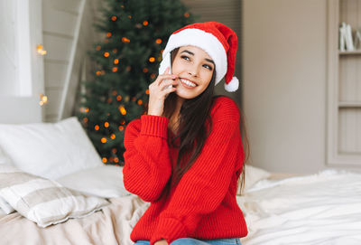 Young asian woman in cozy red knitted sweater and santa hat using mobile in room with christmas tree