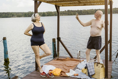 Full length of shirtless senior man photographing woman while standing in gazebo against river