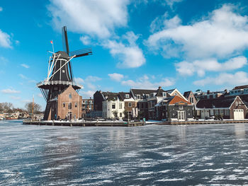 Traditional windmill by buildings against sky and frozen river
