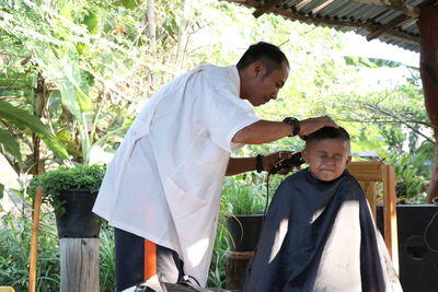 Mature barber trimming scared boy hair at shop against trees