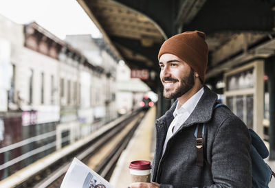 Young man waiting at metro station platform, holding disposable cup