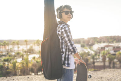 Young man wearing sunglasses standing against sky with guitar on his shoulders