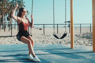 Young woman in sunglasses swinging at beach against clear sky