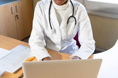 From above crop african american female doctor typing report on laptop while working at table in modern clinic office