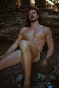 Portrait of shirtless young man sitting by log in lake