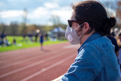 A young man spectator in a blue denim shirt wears sunglasses and face mask at outdoor running track 