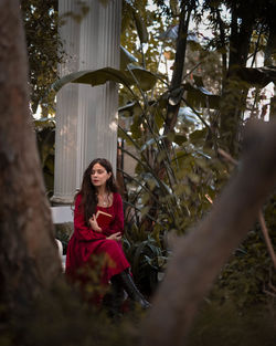 Portrait of young woman daydreaming and sitting in the nature with a book in her long red dress