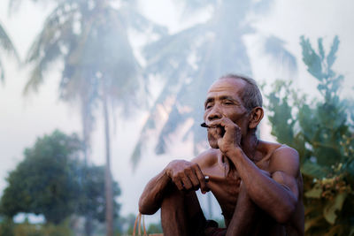 Shirtless senior man smoking cigar against trees