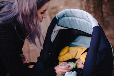 Mother feeding baby son from bottle in baby stroller in park in autumn