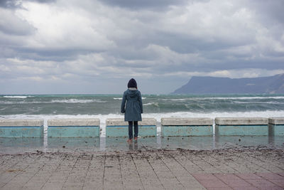 Rear view of woman standing on promenade by sea against cloudy sky