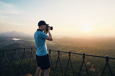 Side view of mid adult man photographing with camera while standing on mountain against sky during sunset