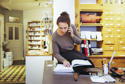 Young saleswoman looking at document while talking on mobile phone sitting in home improvement store