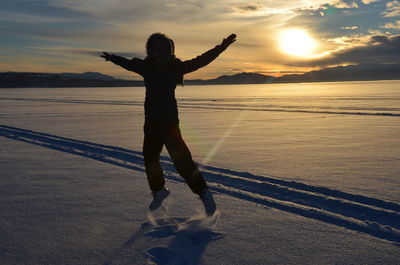 Full length of woman jumping with arms outstretched on snow covered field during sunset